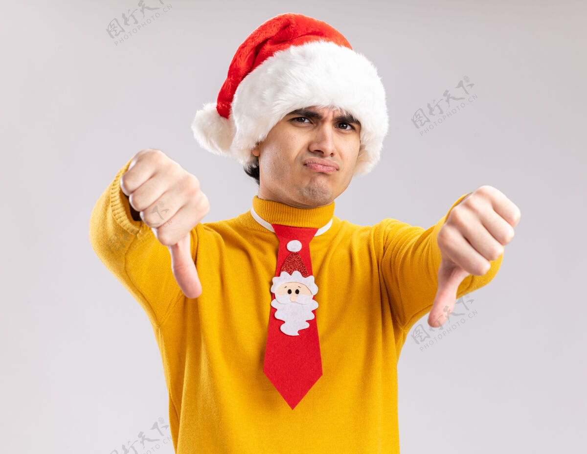 站着一个穿着黄色高领毛衣 戴着圣诞帽 打着滑稽领带的年轻人不高兴地看着摄像机 站在白色背景上竖起大拇指圣诞老人搞笑年轻