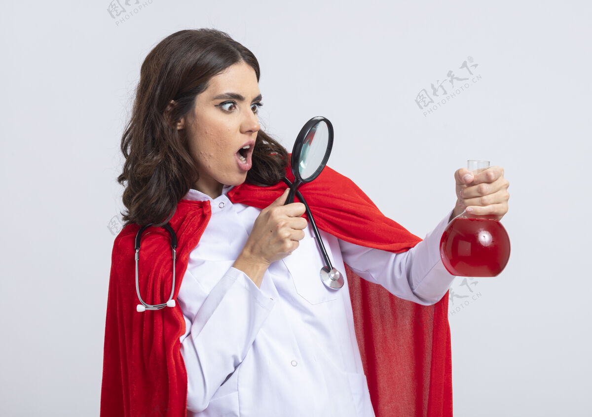 液体一个穿着医生制服 穿着红色斗篷和听诊器的白人超级英雄女孩拿着白色墙上的放大镜 看着玻璃瓶中的红色化学液体超级英雄玻璃医生