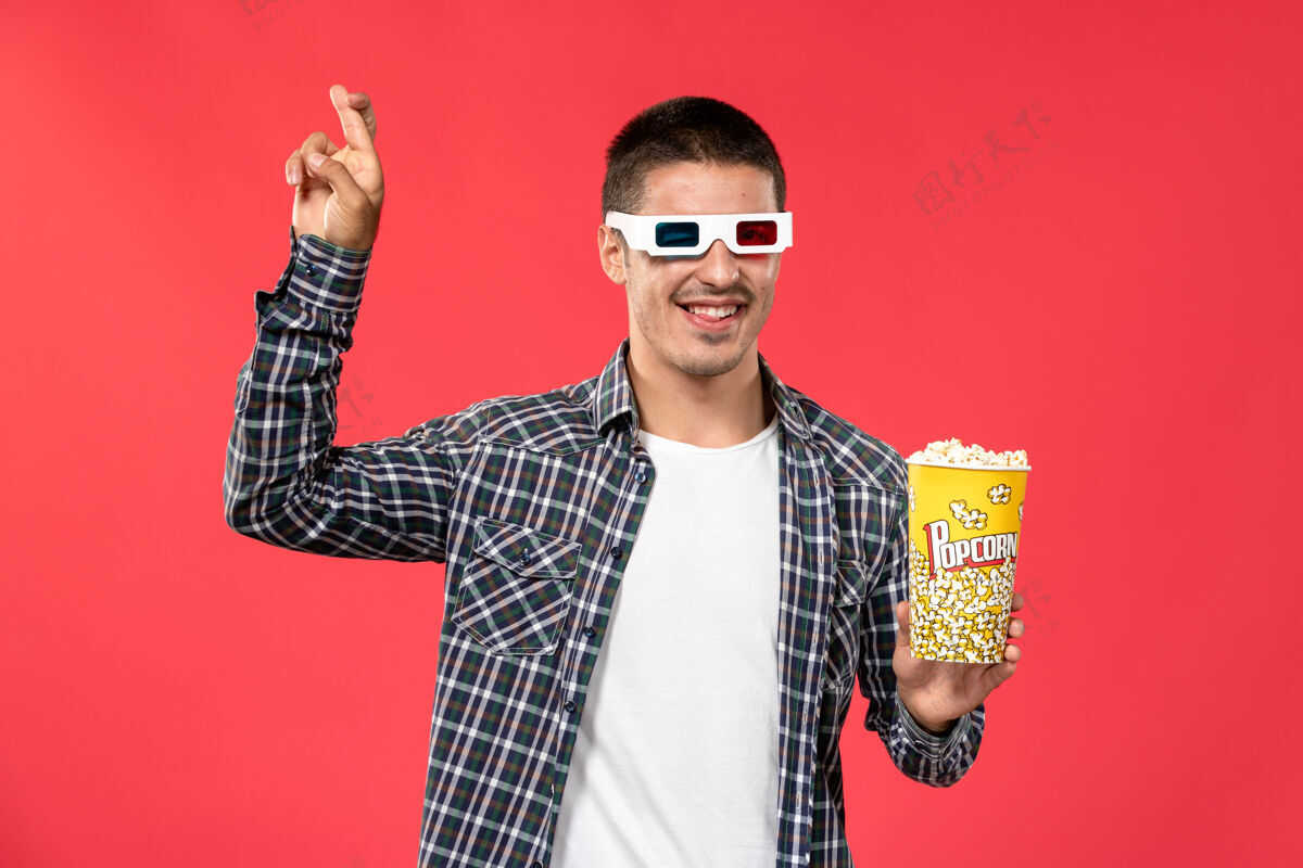 包装正面图年轻男子手持爆米花包 戴着-d太阳镜在淡红墙电影院看电影年轻男性肖像光