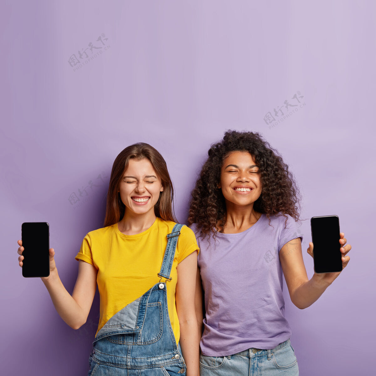 应用程序快乐多样的姐妹们站在一起 展示空白屏幕的手机 拥有积极的外表 为新产品做广告空互联网手持