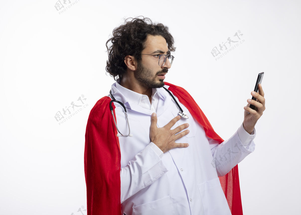 光学震惊的年轻白种人超级英雄戴着眼镜 身穿医生制服 披着红色斗篷 脖子上戴着听诊器 把手放在胸前 看着有复印空间的手机穿着制服听诊器