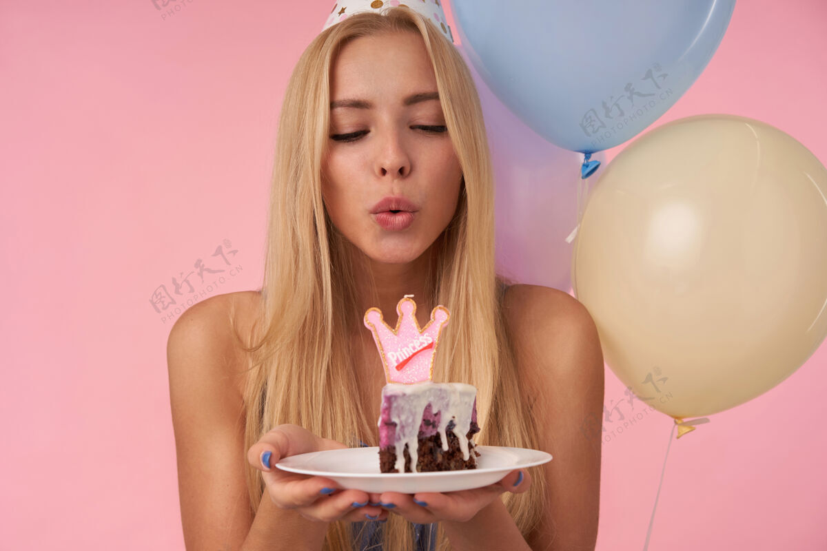 制作积极的年轻长发女性庆祝生日与五彩气球 保持手中的生日蛋糕和吹灭蜡烛 站在粉红色的背景头发裙子五彩