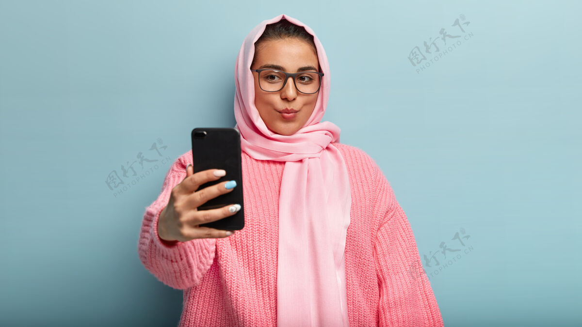 高兴美丽的黑发穆斯林女士录制视频 嘴唇合拢 自拍 捕捉新的面貌 在粉红色围巾和毛衣的姿势 张贴照片在网上的追随者 使室内凉爽的镜头智能手机小玩意毛衣