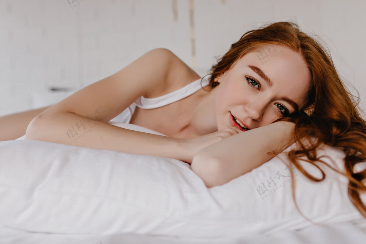 年轻躺在枕头上的快乐的深色妆容的白人女孩睡意朦胧的姜黄色头发的女模特在床上摆姿势床成人红发