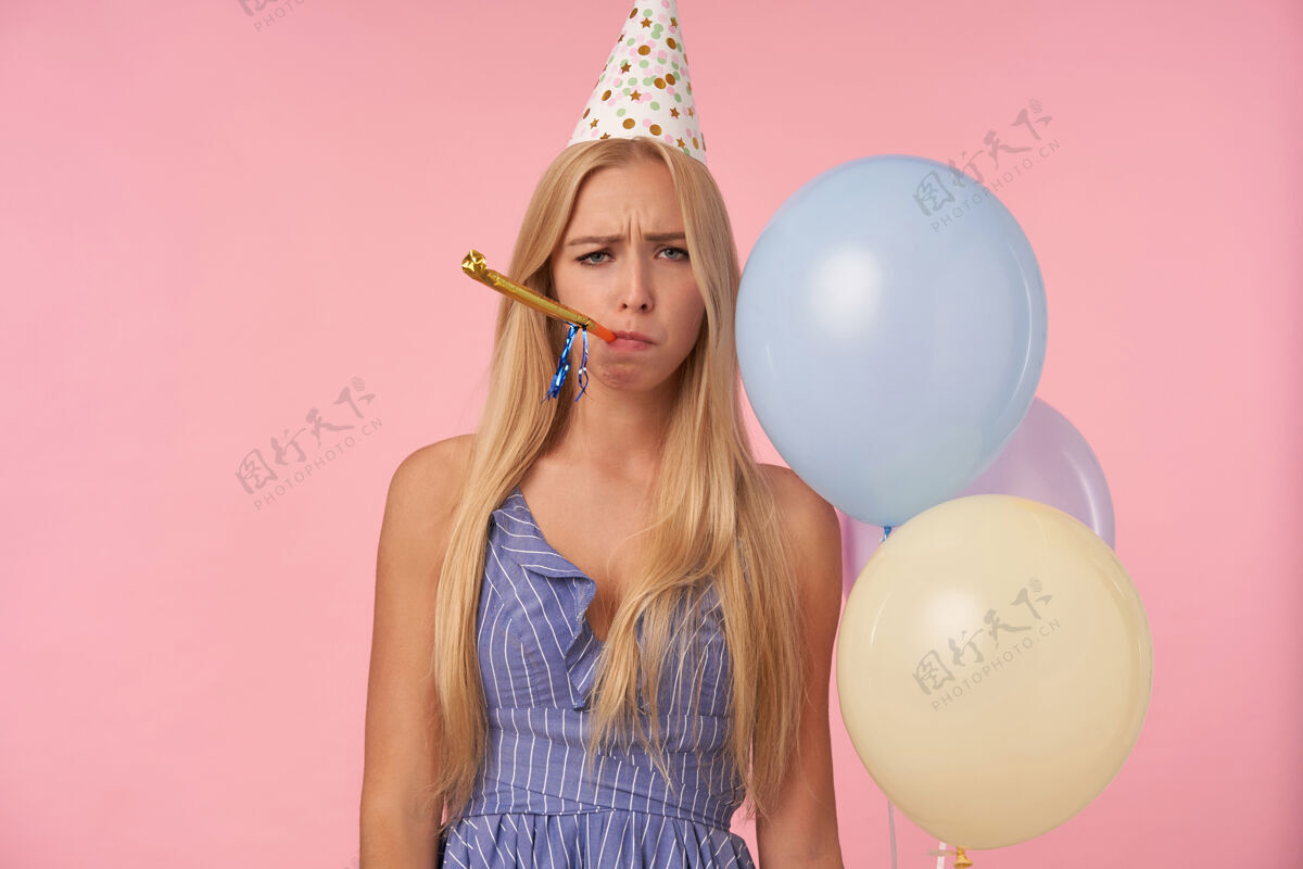 帽子照片中 一位身着蓝色夏装 头戴节日帽的金发女士 在粉色背景下随意吹响派对号角 表情沮丧地看着相机 手里拿着一堆氦气球金发不高兴20多岁