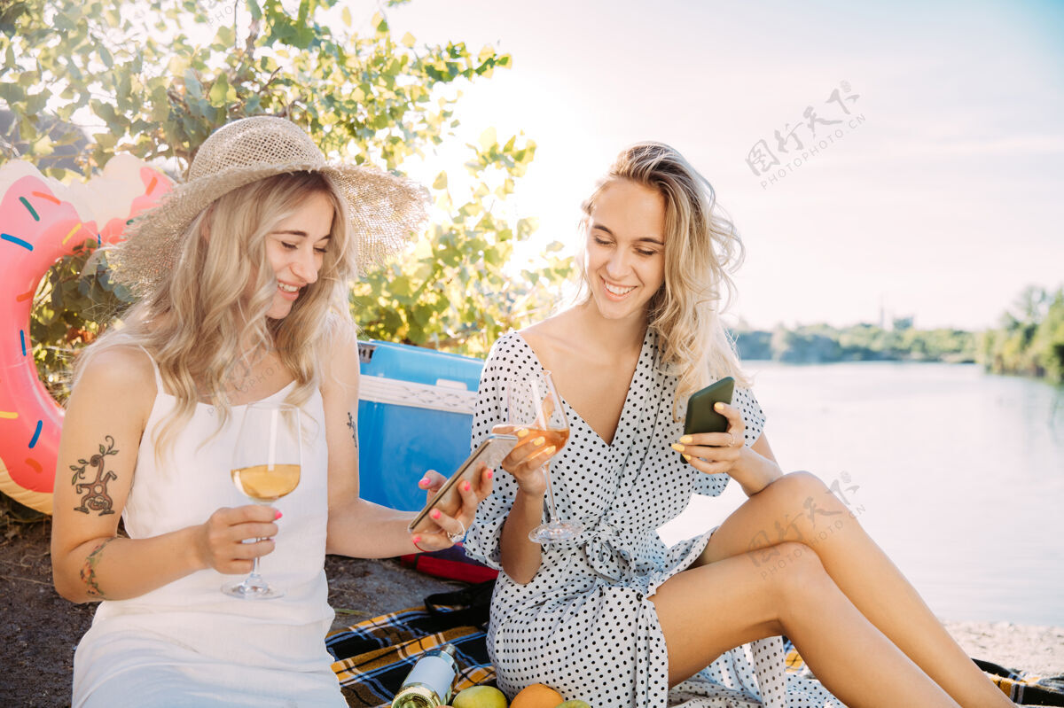 酒年轻的女同性恋夫妇在阳光明媚的日子里在河边玩得很开心女人们一起在大自然中度过时光喝酒 自拍关系的概念 爱情 夏天 周末 蜜月女人乐趣关系