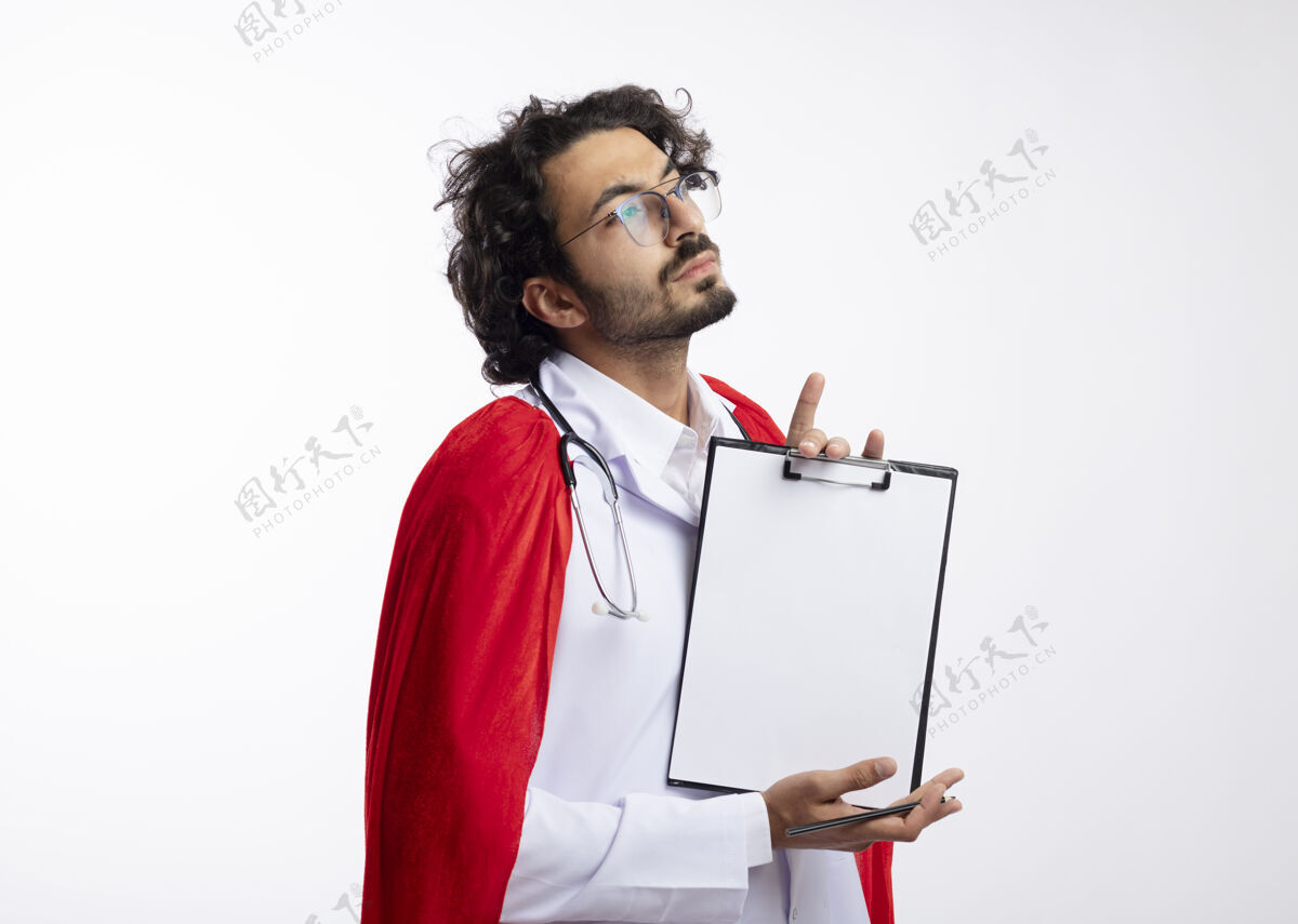 斗篷自信的年轻白种人超级英雄戴着眼镜 身穿医生制服 披着红色斗篷 脖子上戴着听诊器 站在一边 把剪贴板孤立地放在白墙上光学站着男人
