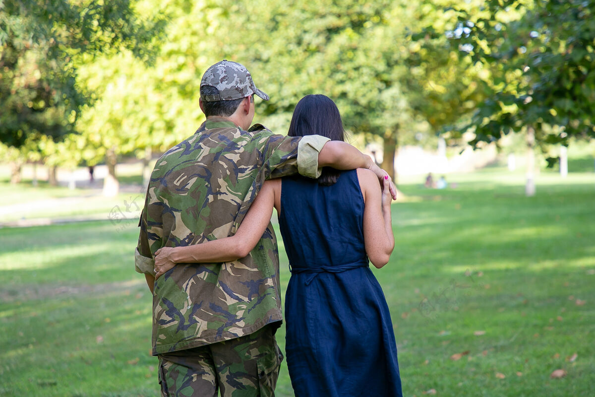 拥抱一对情侣在公园的草坪上拥抱散步的后景男人穿着迷彩服 拥抱着他的妻子 享受着阳光明媚的日子家庭团聚 周末和回家的概念穿着女人士兵