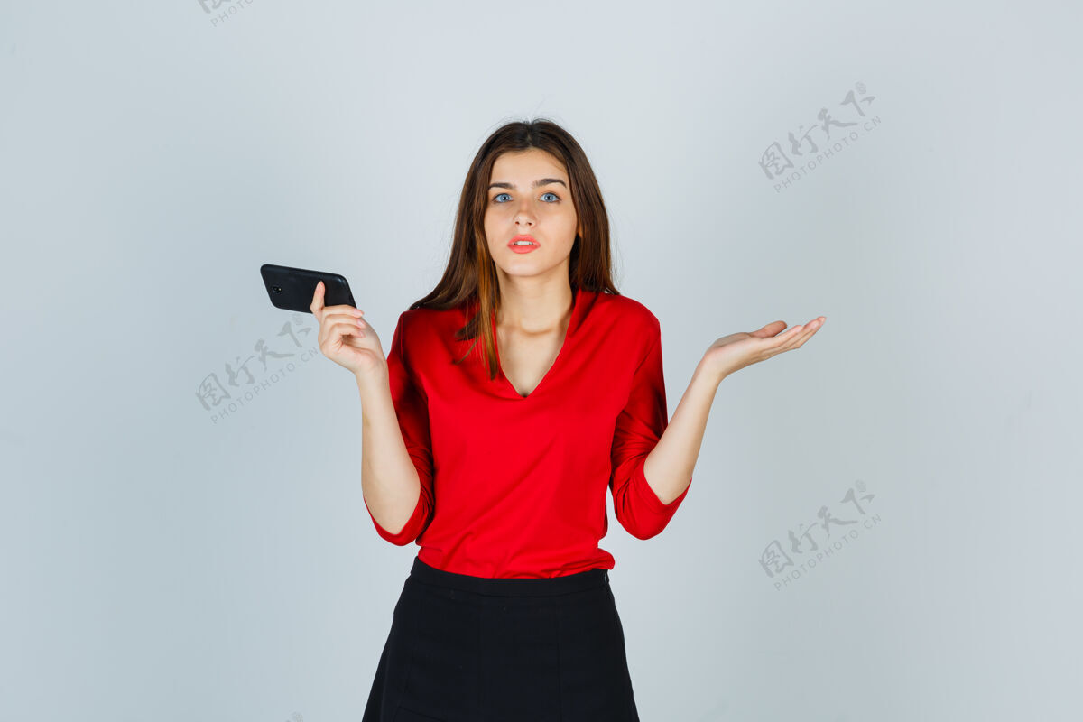 压力一个年轻的女士拿着手机 穿着红衬衫 做着无助的手势问题年轻千禧一代