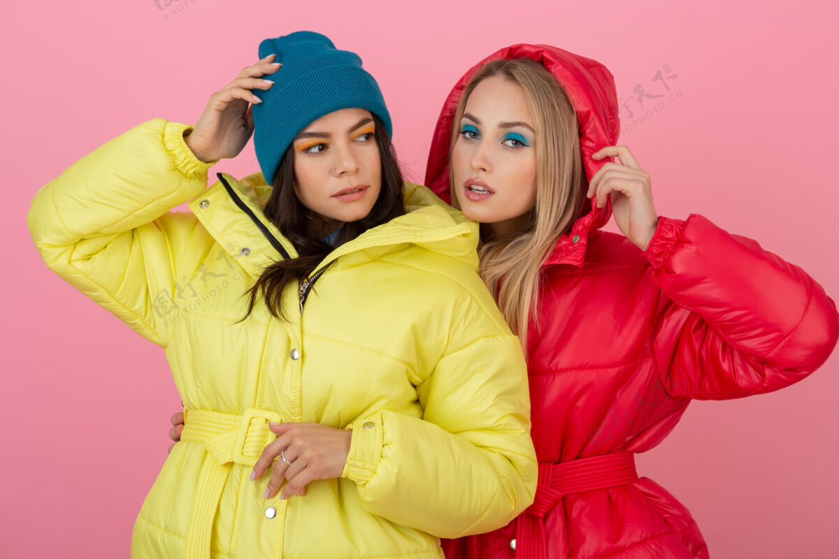 秋季两个迷人的女孩在粉红色的背景下 穿着鲜艳的红色和黄色的彩色冬季羽绒服摆姿势穿着时尚粉色