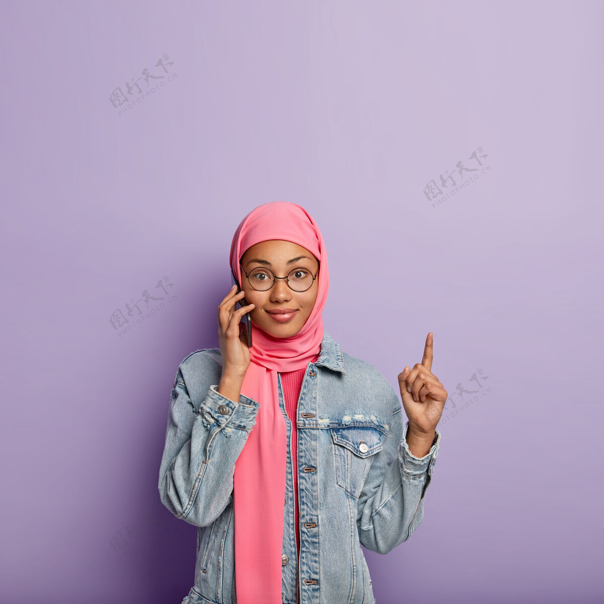 服装漂亮的穆斯林女士和接线员谈论关税 手持现代手机 用食指指着上面的空白处 戴着粉色围巾 隔着紫色的墙电话指向手机