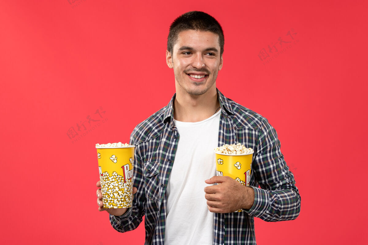微笑正面图年轻男子拿着爆米花包笑着在淡红墙电影院看电影成人爆米花视图