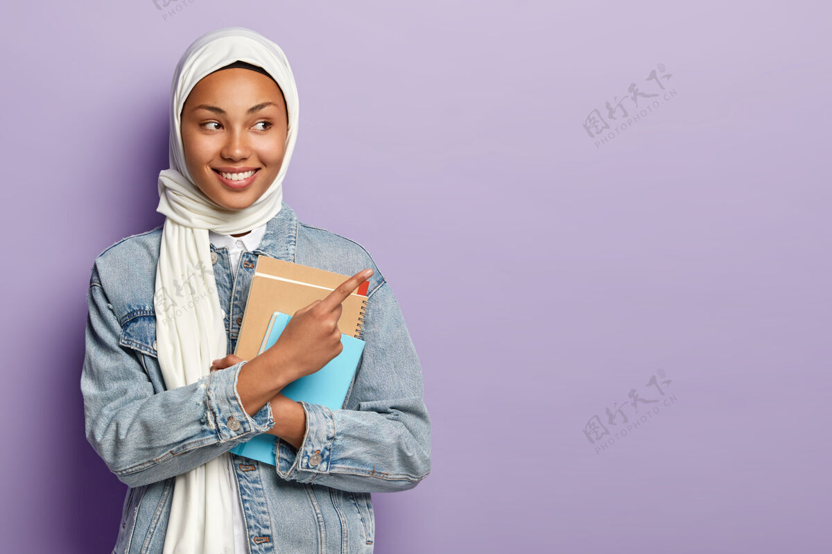 头巾满意的穆斯林学生的孤立镜头显示了可怕的广告 在右上角点 穿着白色面纱和牛仔夹克情感指向笔记本