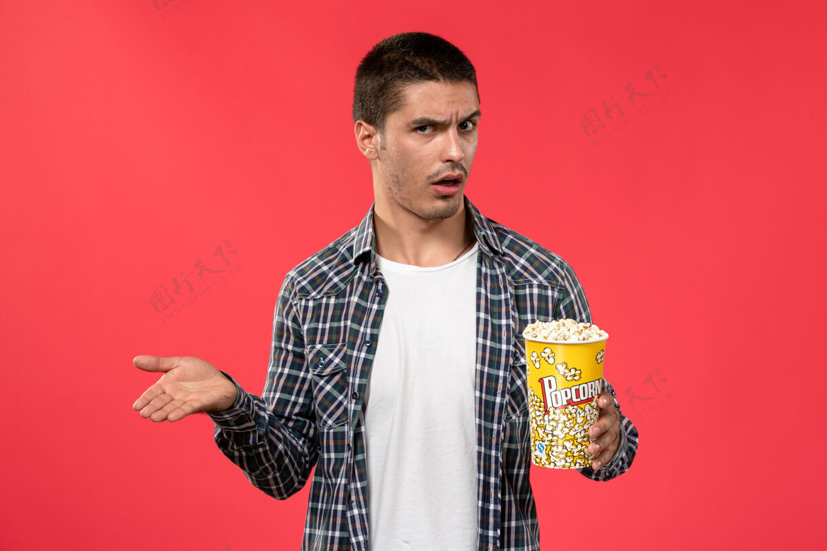 电影正面图年轻男子拿着爆米花包 表情困惑地站在淡红色的墙上看男性电影男人肖像包装