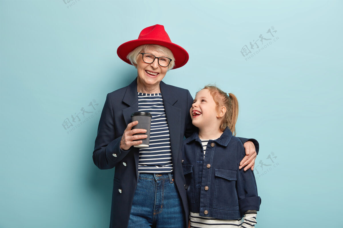 童年快乐时尚的老奶奶和小可爱的孩子在一起度过业余时间 喝外卖咖啡 拥抱 有友好的关系 穿牛仔服 站在蓝色的墙上积极孙女老年人