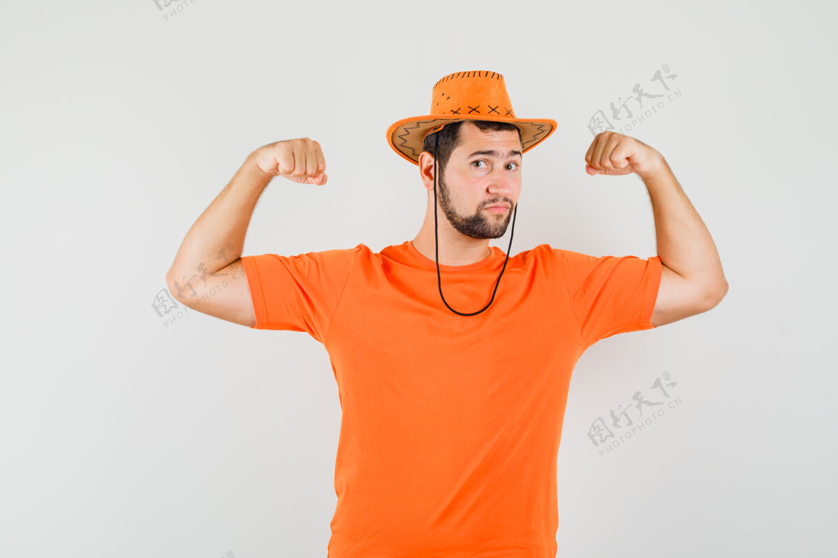 模特身穿橙色t恤 戴着帽子 看上去很强壮的年轻人展示手臂肌肉前视图帽子手势成人