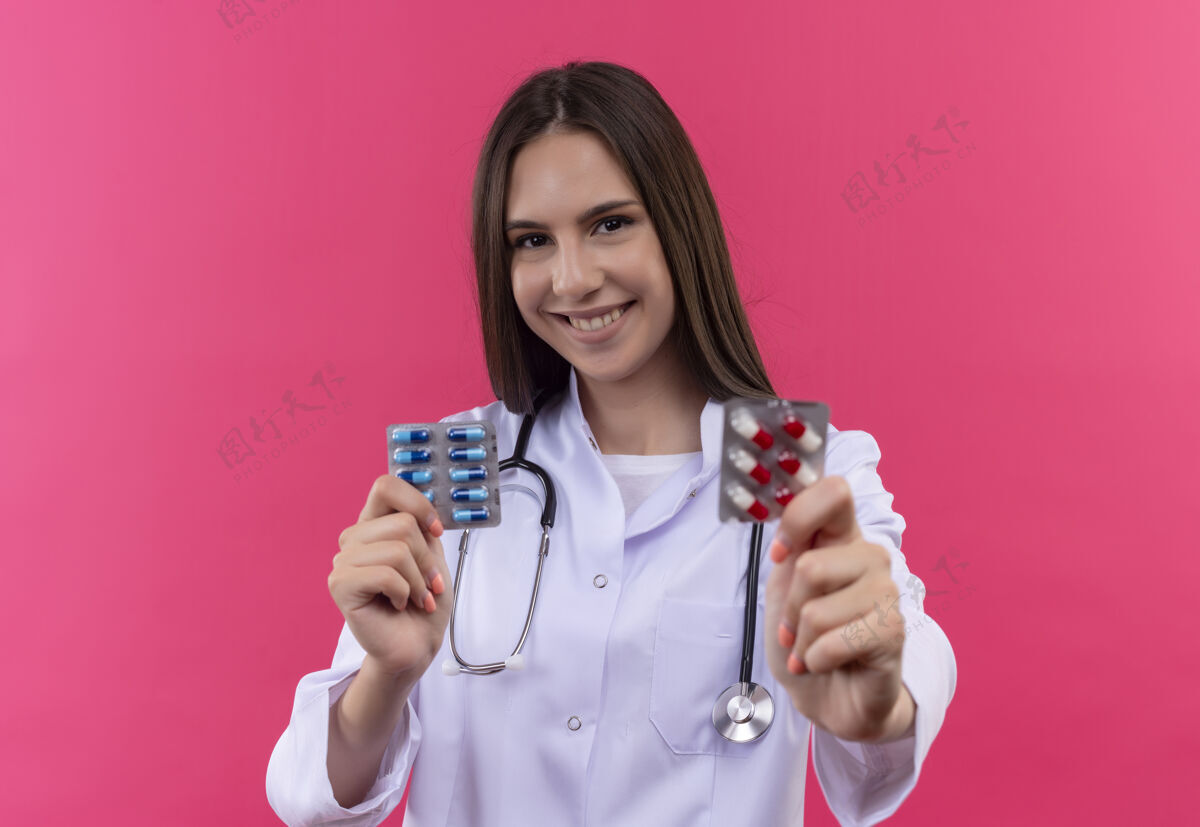 抱着微笑的年轻医生女孩穿着听诊器医用长袍 拿着药片 背景是孤立的粉红色微笑药片粉红