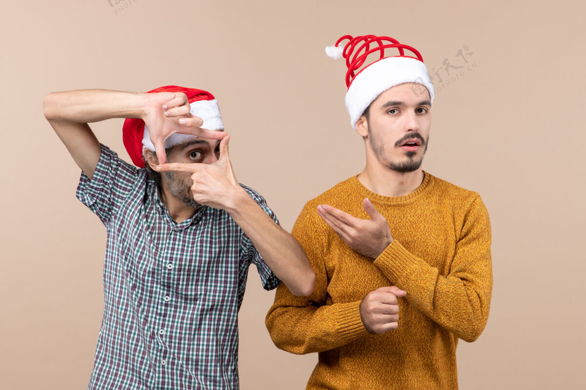 手前视图两个戴着圣诞帽的家伙一个用手做摄像机标志 另一个用米色孤立的背景展示他两个伙计们圣诞老人前面