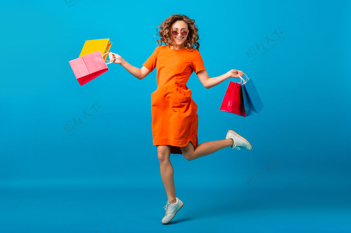 女士迷人的快乐微笑时尚女性购物狂穿着橙色时髦的超大号连衣裙在蓝色工作室背景上拿着购物袋跳跃零售休闲销售