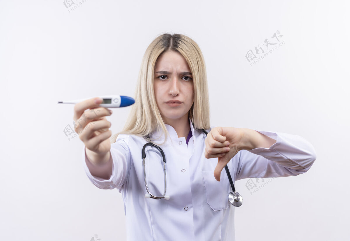 按住严格的医生年轻的金发女孩戴着听诊器和医用长袍拿着温度计对着摄像机她拇指朝下对着孤立的白色背景穿着长袍医疗