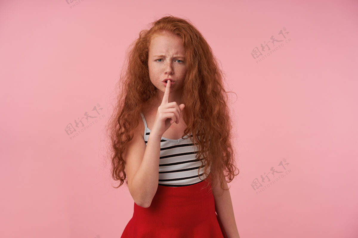 优雅一张严肃的红头发女孩的摄影棚照片 她留着长长的卷发 穿着红色裙子和条纹上衣 把手指放在嘴唇上 对着镜头皱眉 要求保持沉默 站在粉色背景上雀斑手势长