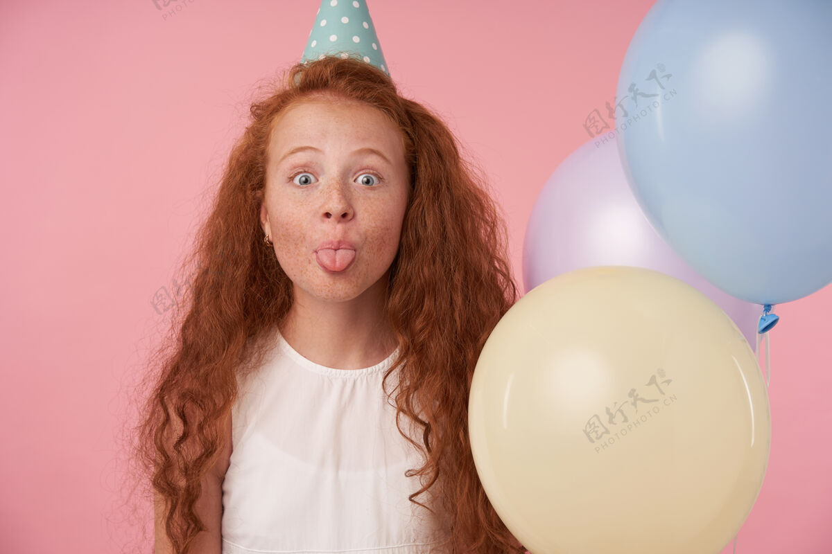 舌头一个有趣的红头发女孩的特写镜头 卷发 白色连衣裙 戴着生日帽 庆祝着什么 开心地看着镜头 露出舌头在粉色背景上摆出彩色气球的姿势卷曲头发衣服