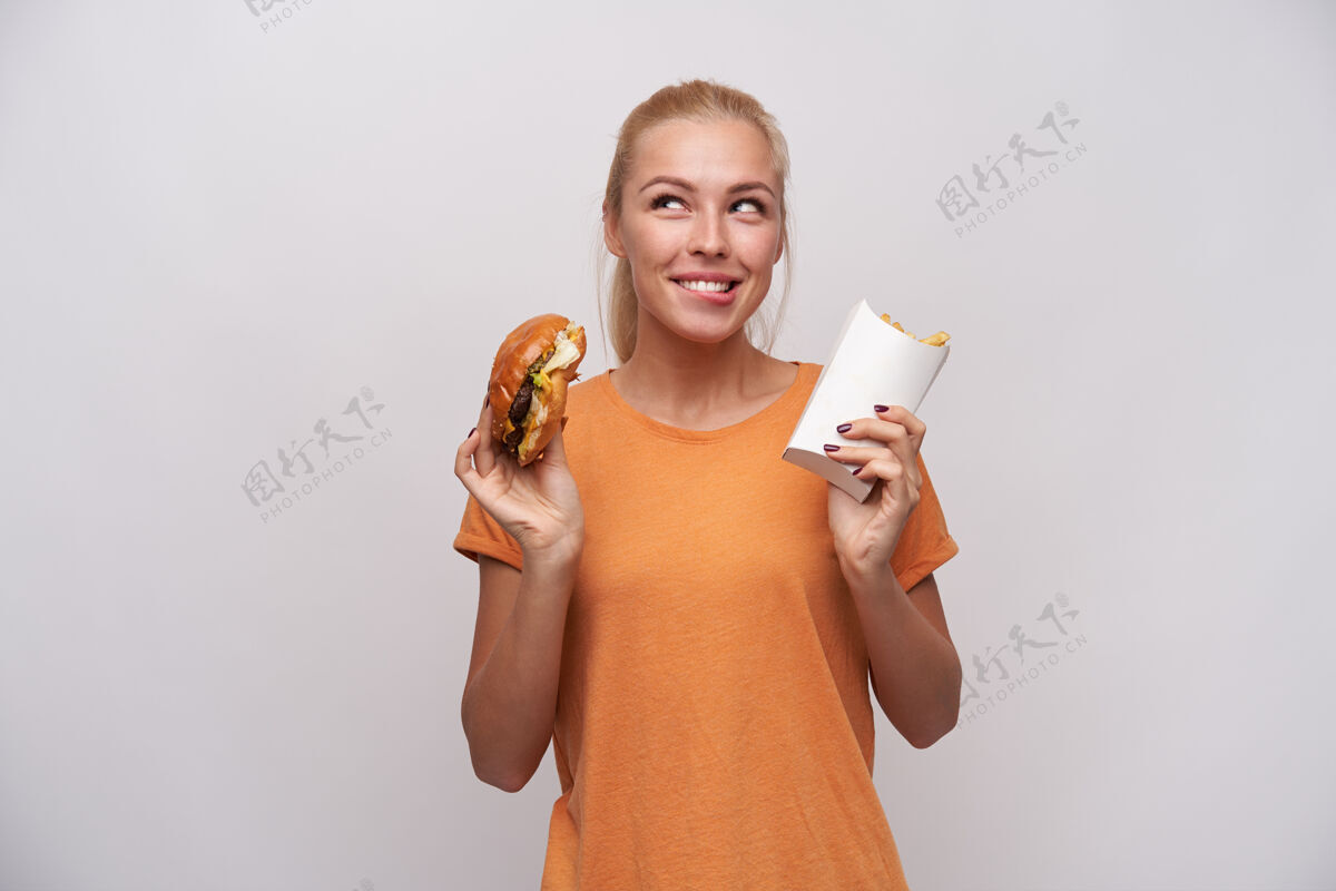 勃艮第摄影棚拍摄了一位年轻的金发女郎 她留着随意的发型 手里拿着汉堡包和炸薯条 兴高采烈地看着一旁 咬着内衣 预感美味的晚餐饮食积极饥饿