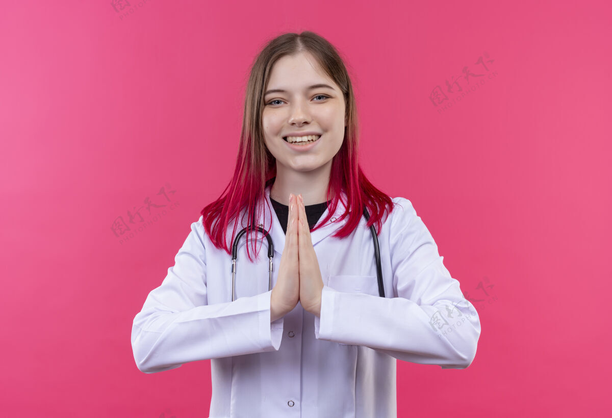 微笑微笑的年轻医生女孩穿着听诊器医用长袍 在粉红色的孤立背景上显示祈祷的手势手势医疗年轻
