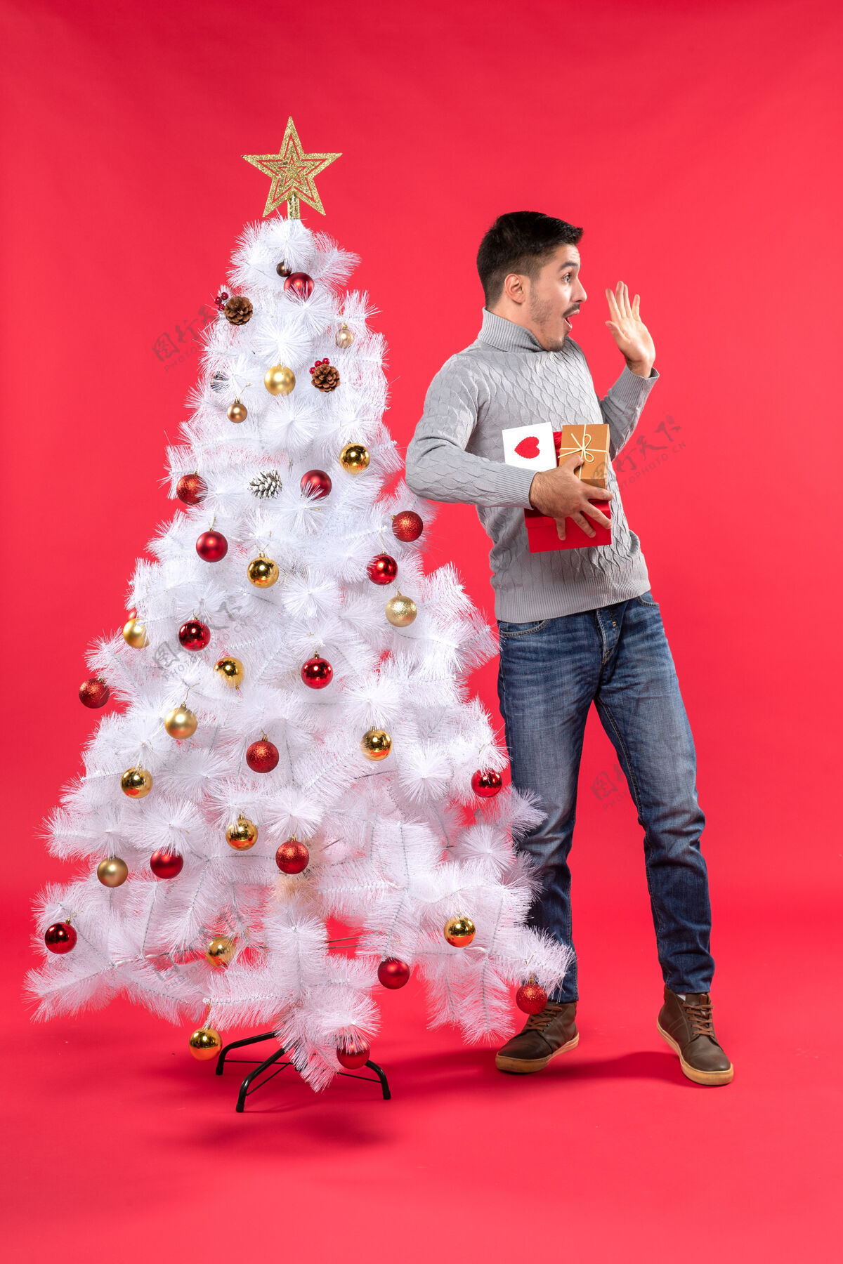 装束年轻帅气的大人站在装饰好的白色圣诞树旁 手里拿着礼物红色微笑成人