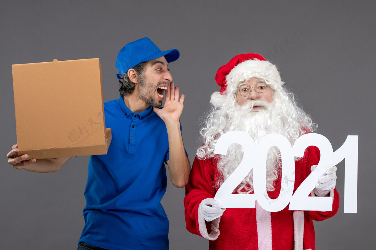 服装圣诞老人的正面图 男信使拿着食物盒 站在灰色的墙上盒子快乐庆祝