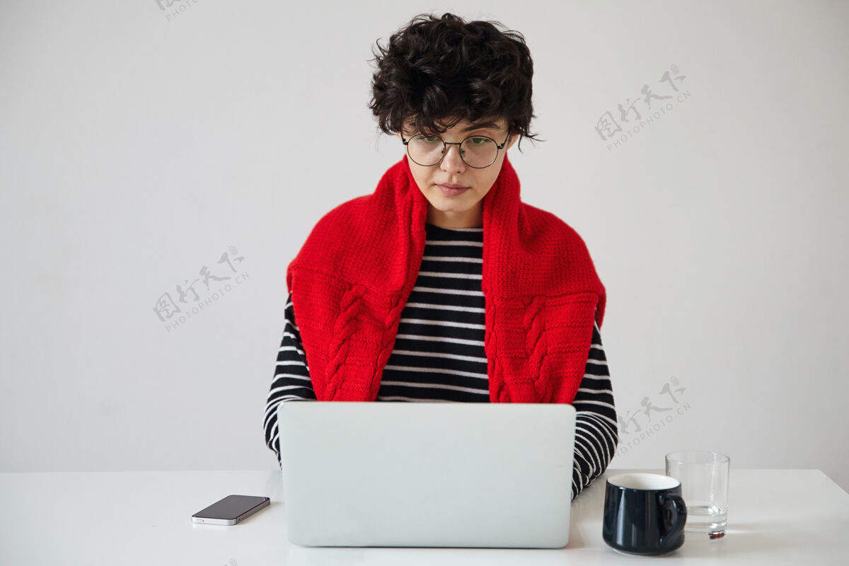 工作严肃的年轻黑发卷发女士 短发时髦 戴着眼镜 用现代笔记本电脑在白色背景下工作 凝神看着屏幕羊毛室内时尚