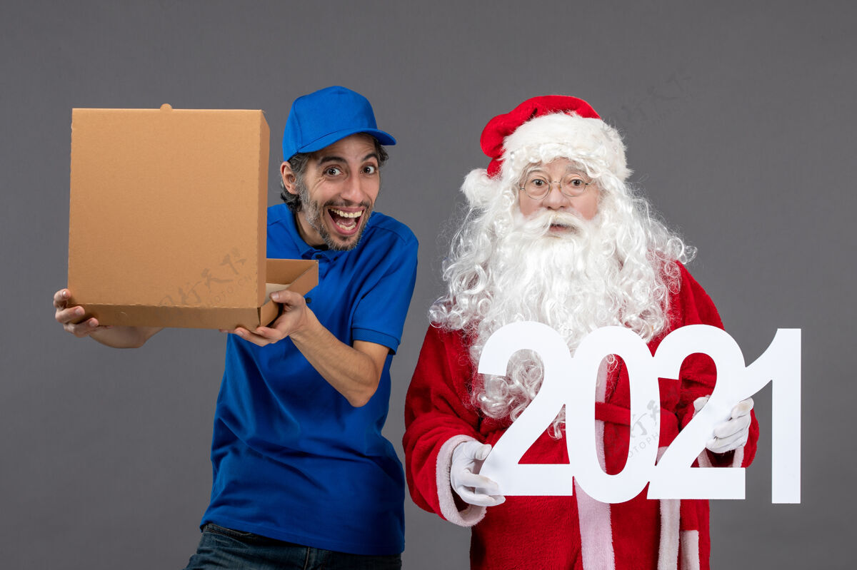 服装圣诞老人的正面图 男信使拿着食物盒 在灰色的墙上写字快乐前面举行