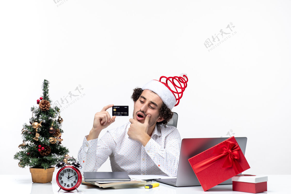 圣诞老人年轻忙碌的商务人士戴着圣诞老人的帽子 拿着他的银行卡 在白色背景的办公室里仔细地头脑风暴办公室商务人士人