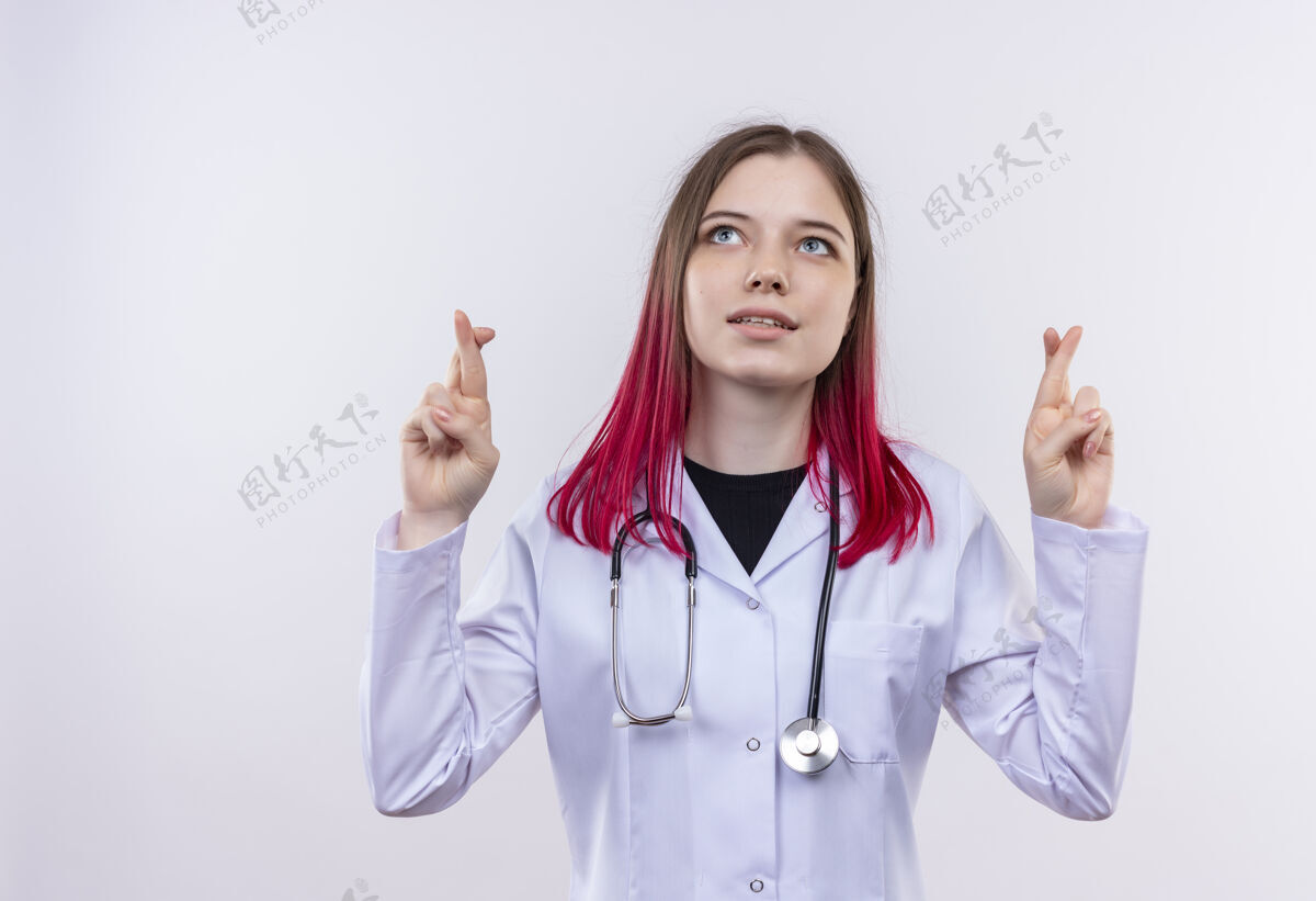 医疗看着年轻的医生女孩穿着听诊器医用长袍交叉手指孤立的白色背景看白色向上