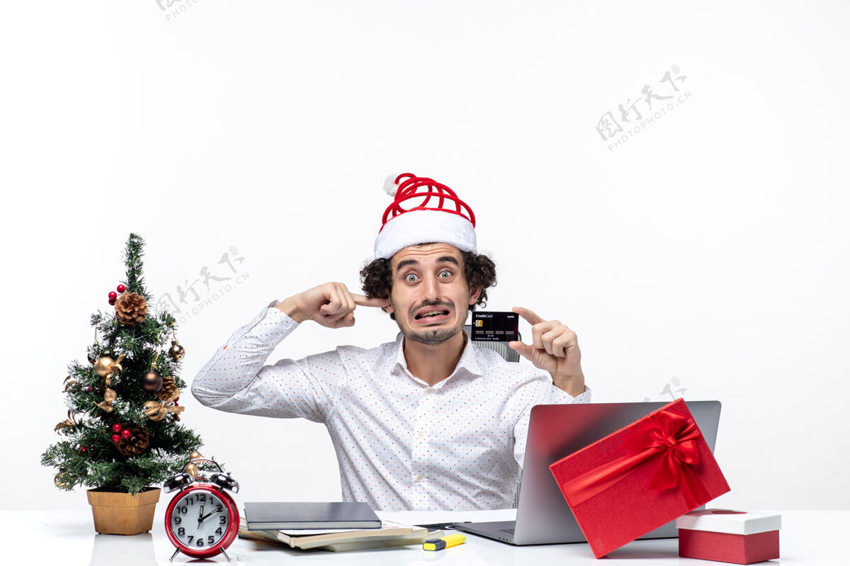 耳朵圣诞节心情与年轻的胡子紧张的商人与圣诞老人的帽子举行银行卡和关闭他的耳朵在办公室圣诞老人办公室生意人