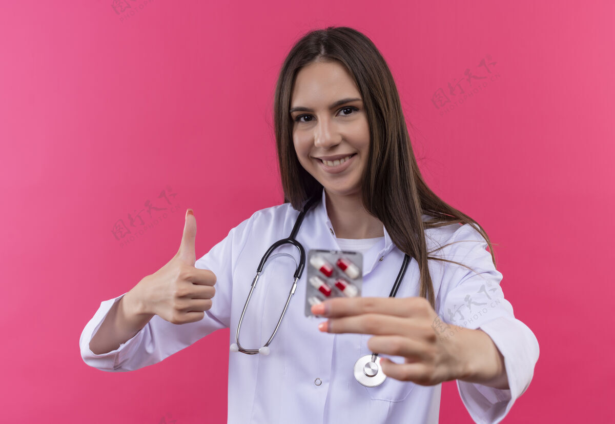 医疗微笑着的年轻医生女孩穿着听诊器医用长袍拿着药片 她的拇指在孤立的粉红色背景上向上粉红向上年轻