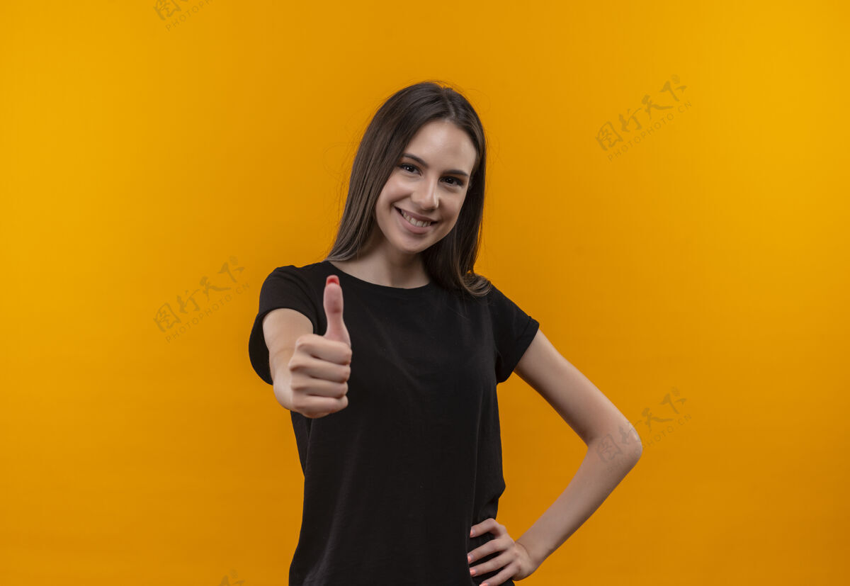 臀部微笑的年轻白人女孩穿着黑色t恤 大拇指向上 手放在臀部 背景是孤立的橙色微笑拇指黑色