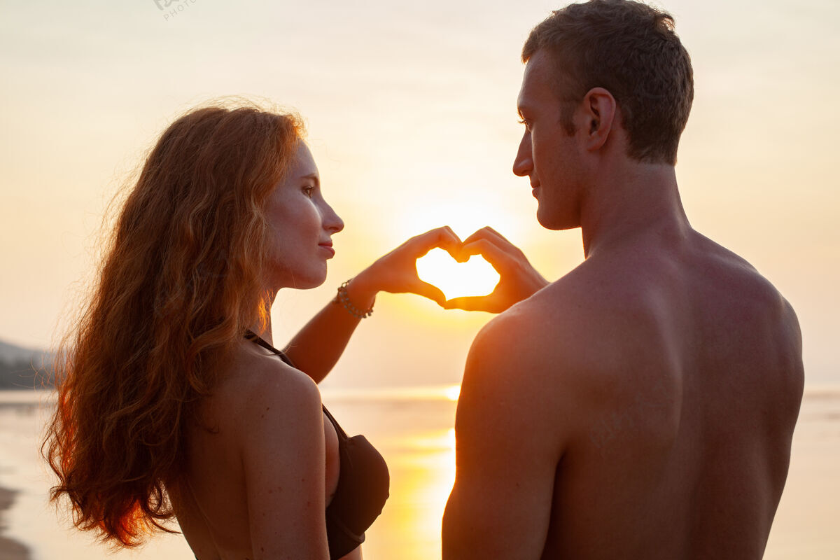 在一起年轻性感浪漫的情侣在夏天的沙滩上快乐地在一起 穿着泳衣在日落时露出心形标志约会情人看
