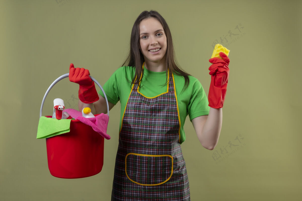 海绵高兴的清洁年轻女孩穿着制服 戴着红色手套 拿着清洁工具和海绵 背景是孤立的绿色手握绿色红色