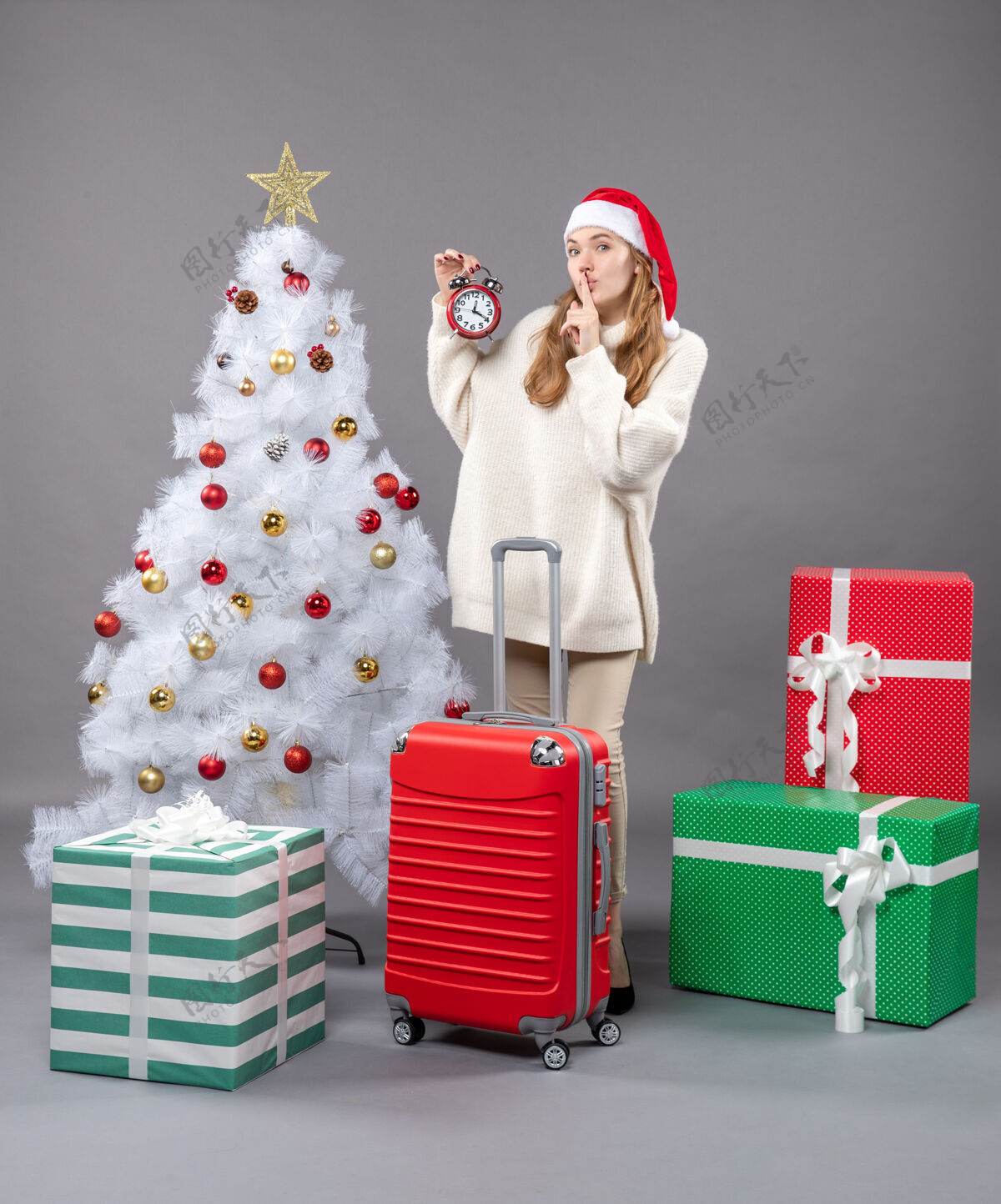 礼物正面图金发女孩戴着圣诞帽手持红色闹钟显示嘘手势假期手提箱帽子