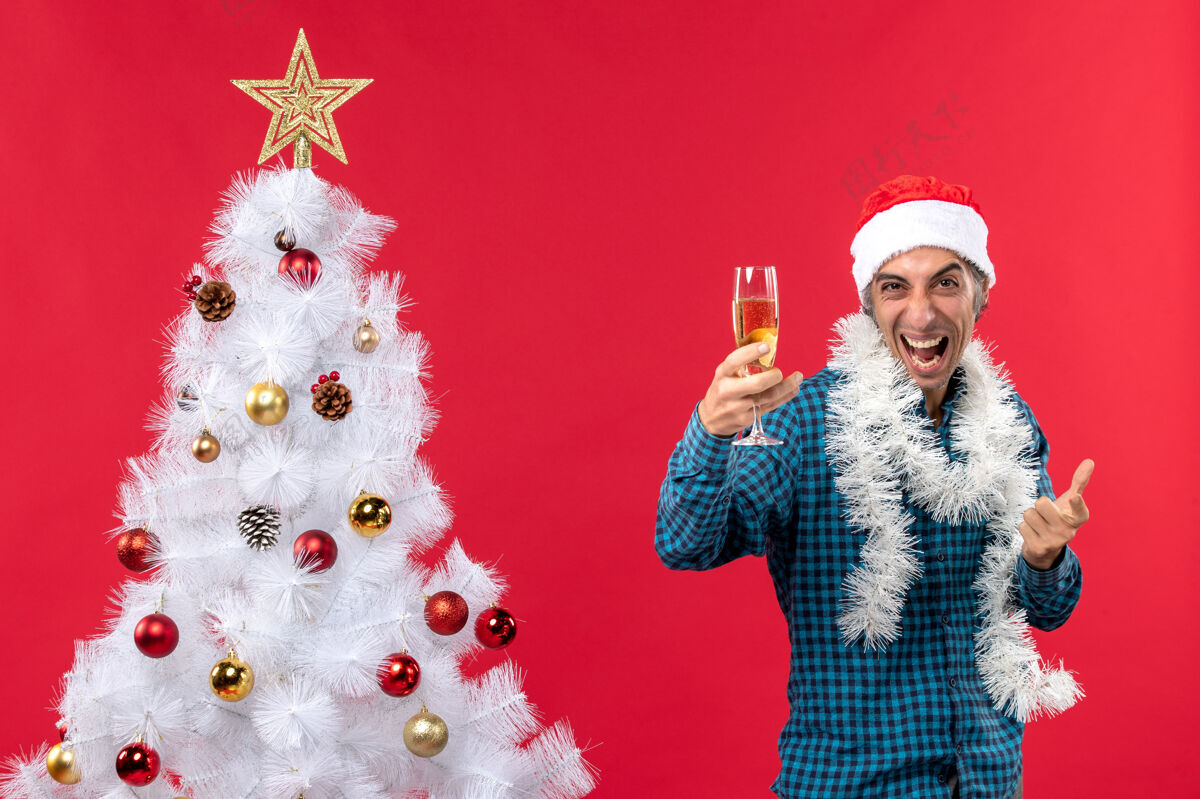 年轻人圣诞节心情与情绪疯狂的年轻人与圣诞树附近拿着一杯葡萄酒的蓝色条纹衬衫圣诞老人帽子举行聚会小伙子