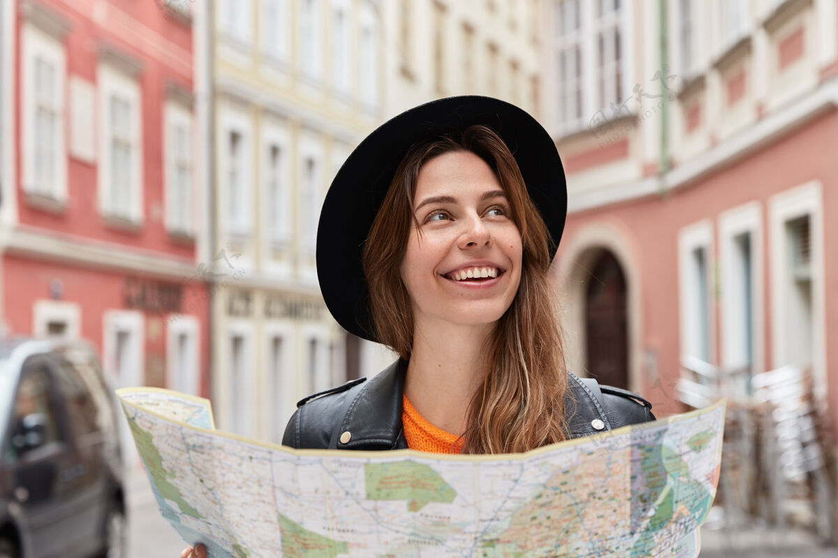 女士快乐乐观的女性旅游者在地图上找到了自己的位置 在夏天的旅行中漫步在市中心 戴着时髦的黑帽子 在城市的背景下摆姿势远足城市城市
