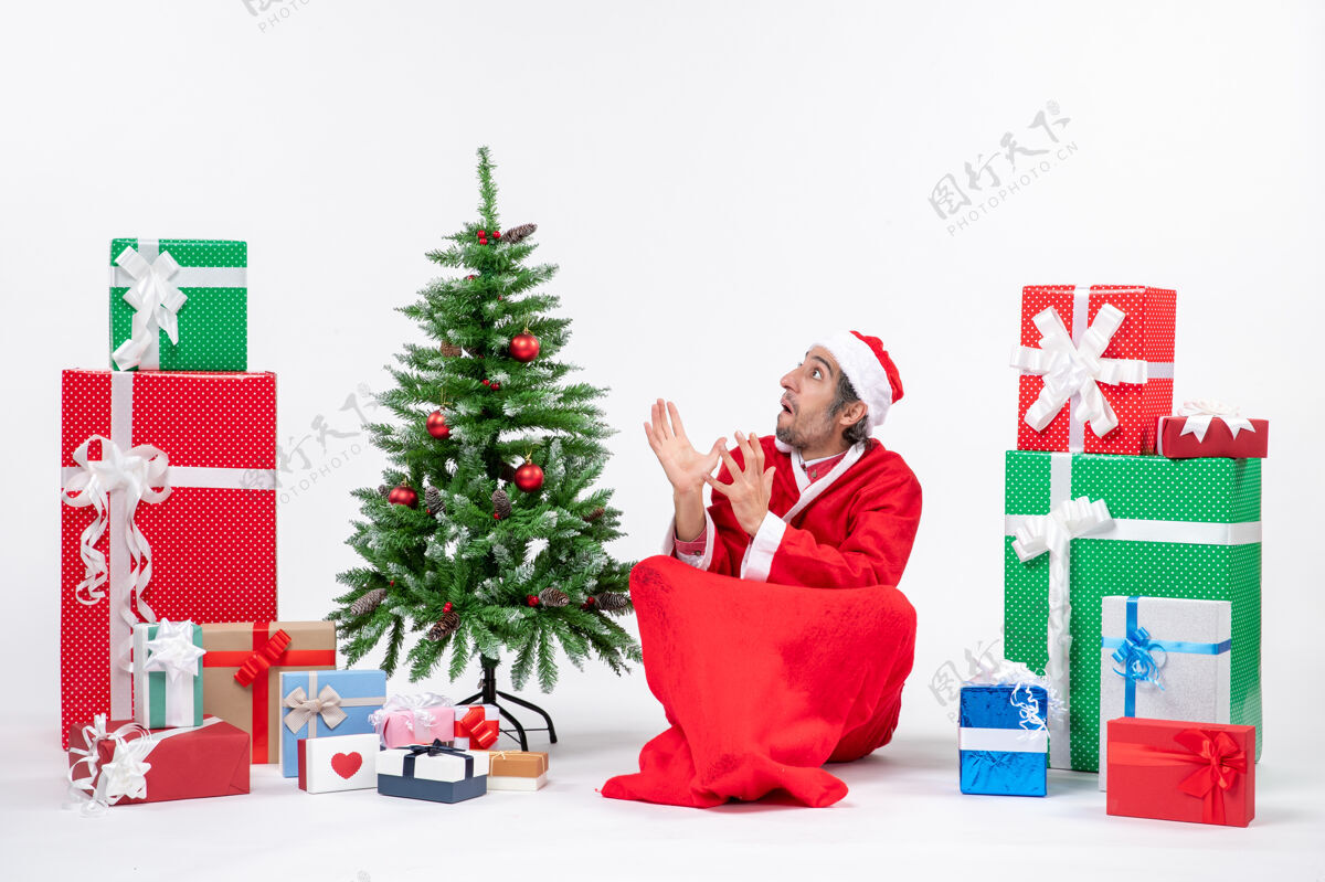 装饰被吓坏了的年轻人打扮成圣诞老人 拿着礼物和装饰好的圣诞树坐在白色背景的地上圣诞礼物圣诞树