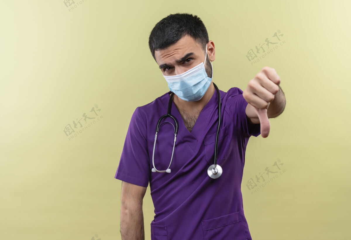 衣服悲伤的年轻男医生穿着紫色的外科医生服装 戴着医用听诊器 把大拇指放在孤立的绿色背景上年轻绿色男性