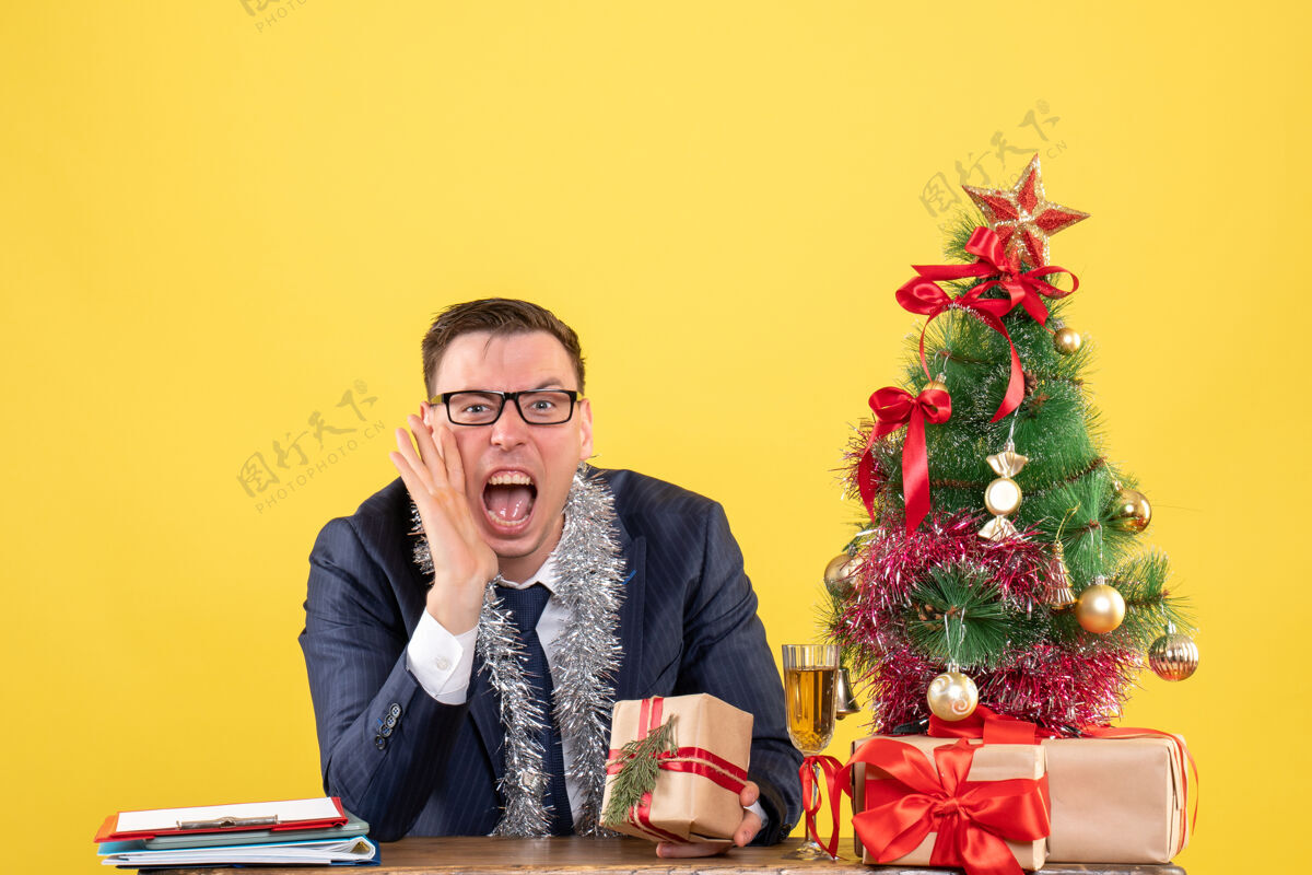 男人一个年轻人对着坐在圣诞树旁的桌子上的人大喊大叫 并把礼物穿上黄色坐着办公室树