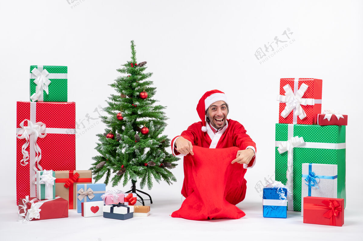 人笑容满面的年轻人打扮成圣诞老人 拿着礼物和装饰好的圣诞树坐在白色背景的地上圣诞老人坐礼物