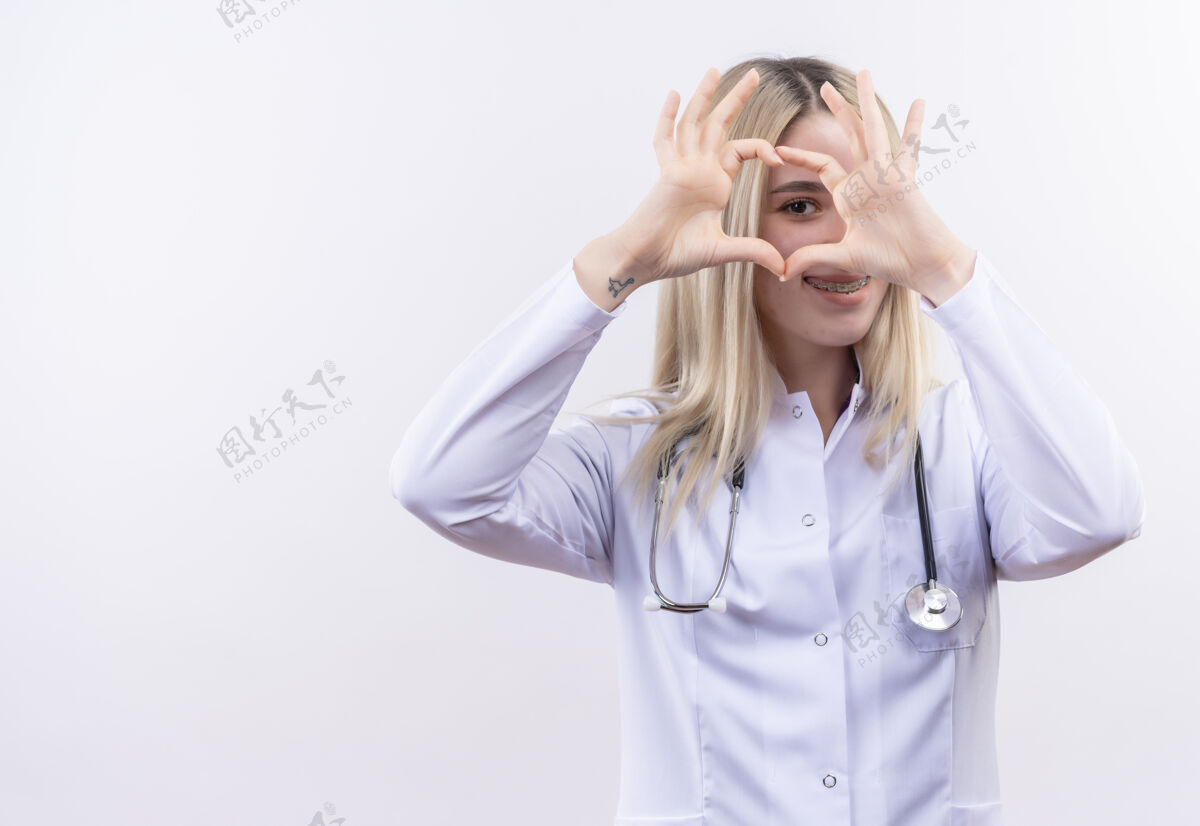 年轻微笑的医生 年轻的金发女孩 戴着听诊器 穿着医用长袍 戴着牙套 在孤立的白色背景上展示着心脏的姿态长袍穿着白人