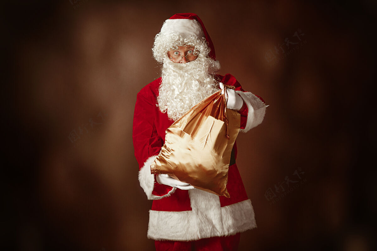 圣诞老人穿着圣诞老人服装的人的肖像-一个豪华的白胡子 圣诞老人的帽子和红色服装在红色工作室的背景与礼物传统季节父亲