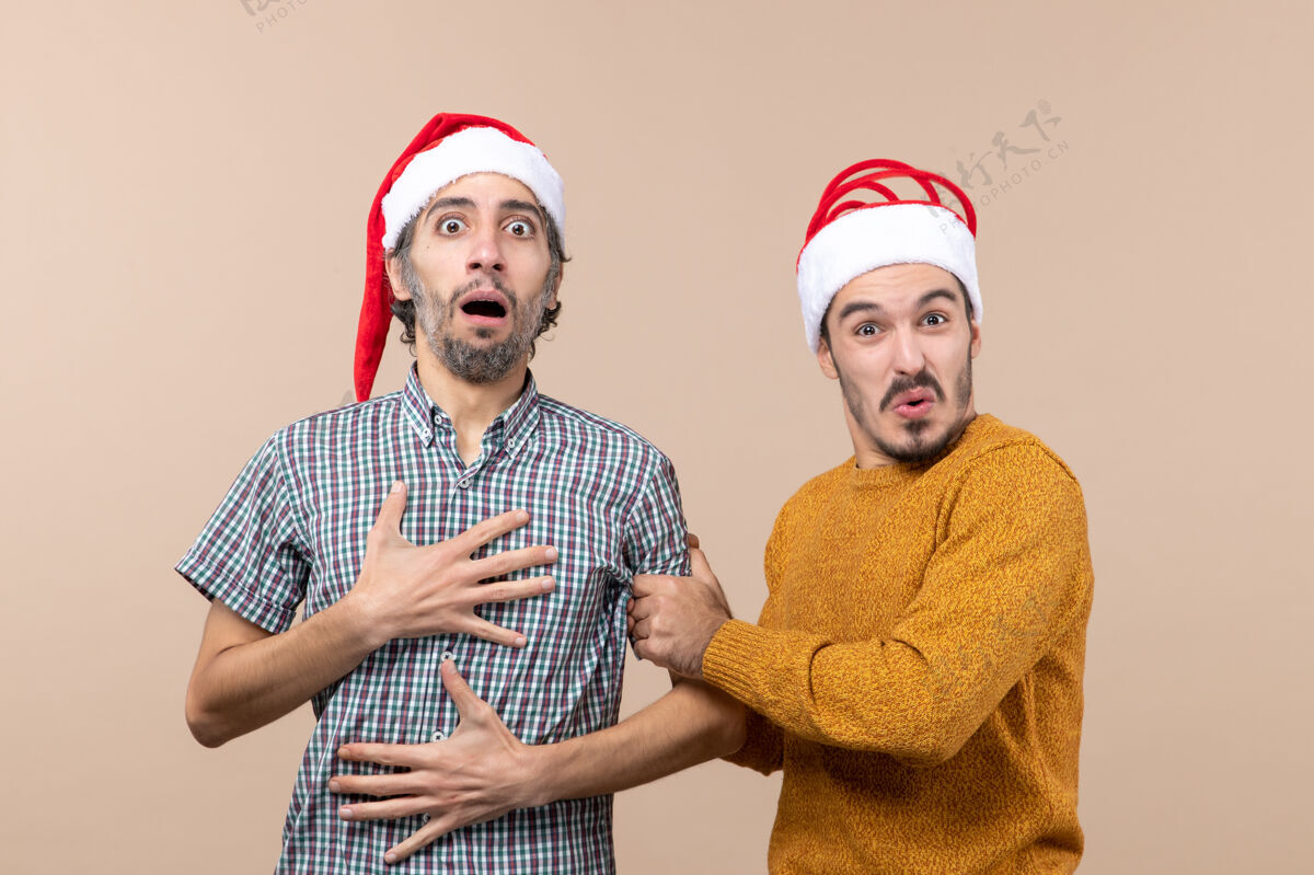 伙计们正面图两个戴着圣诞帽的好奇的家伙 一只手放在他胸前 背景是米色的微笑帽子圣诞老人