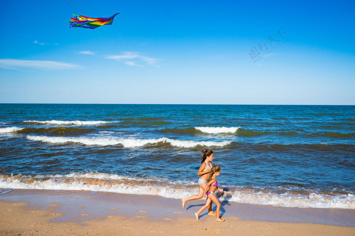 沙两个快乐的小女孩带着风筝在海边的沙滩上奔跑阳光明媚温暖的夏日活跃的儿童游戏概念复制空间暑假湖万里无云